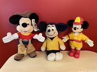 Photo showing three vintage Mickey Mouse plush toys: Western Mickey, Knickerbocker Mickey, and Firefighter Mickey, displayed against a red background.