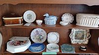 Shelf display showing multiple assorted decorative china and ceramics including plates, bowls, a butter dish, and a covered tureen with floral and scenic patterns.