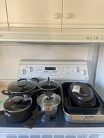 Full view of the four black pots with glass lids arranged on stovetop alongside variety of oven pans, including black speckled and metal pans.