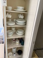 Full view of cabinet shelves containing Corelle dinnerware, bowls, mugs, and additional glassware including mugs and drinking glasses.