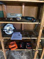 Photo showing shelf holding various power tools and accessories, including a circular saw with blade, an orange jigsaw, a measuring tape, trowel, and a set of drill bits.
