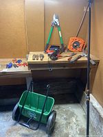 Overview of assorted garden equipment including hose reel, pruners, spreader cart, hand tools on workbench.