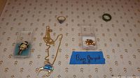 Photo showing a collection of jewelry including a gold-filled pin by Bon Boyd labeled with blue tape, a jade ring, a gold-tone ring with a blue gemstone, a gold chain, and a pair of square earrings with black and gold accents.