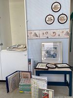 Photo shows corner with blue and white bench, multiple framed needle point and cross stitch artworks, crafts storage boxes, and fabrics.