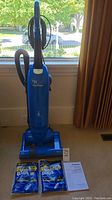 Full view of blue Panasonic upright vacuum cleaner standing upright next to window with two packs of vacuum bags and user manual in front.