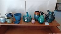 Wide view of nine assorted ceramic and glass decorative items including pitchers, vases, and a lidded candy dish on a wooden shelf