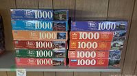 Photo showing stacked boxes of ten 1000-piece puzzles from MB and Cardinal with various scenic images on box art.