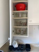 Tall kitchen cabinet with seven kitchen items: red plastic mixing bowl on top shelf, clear glass mixing bowl on second shelf, and three Pyrex measuring cups of various sizes on bottom two shelves. Below counter are muffin pans and white mixer.