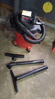Red and black Craftsman wet/dry vacuum with attached black hose and visible power cord, standing on orange wheels. Several black attachments placed on the floor near the vacuum including a floor nozzle and extension tubes.