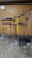 Wide view of eight garden tools and snow shovels leaning against a wall on the floor beneath wooden shelves with small items.