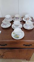 Full view of all 7 bone china tea cups and saucers with different floral designs on wooden surface.