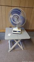 Full view of vintage Super brand electric fan sitting on wood folding table, showing fan blades and table surface.