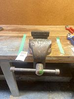 Front top-down view of a well-used heavy metal bench vice attached to a wooden bench, showing the T-handle and vice jaws.