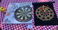 Shows the magnetic dartboard box with darts and a foldable dartboard pouch on a red and white checkered table.