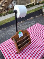 Photo showing the wooden magazine holder in front of the black metal toilet paper stand with a large roll on it, both placed on a red and white checkered tablecloth outdoors.