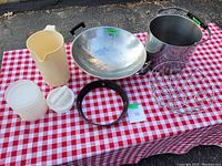 Photo of all cookware and kitchen items on a red and white checkered tablecloth showing wok, stock pot, juice jug, fruit bowl, plastic containers, and burger press