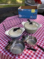 Set of enamel pots, aluminum saucepans, and roasting rack displayed on red and white checkered tablecloth.