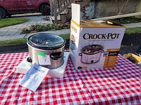 Front and side view of Crock-Pot slow cooker with glass lid, original box packaged nearby, electric cord visible, placed on red and white checkered tablecloth.