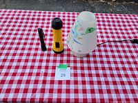 Photo showing two flashlights (one black, small with clip, one yellow) and one children's bedtime lamp with translucent shade decorated with colorful shapes, all placed on a red and white checkered tablecloth.