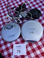Two Sony Discman CD Walkman portable players, silver and white models, with wires and headphones on red and white checkered background.