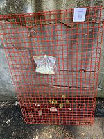 Red metal grid shelf leaning against a concrete wall with a plastic bag of mounting screws attached to it.