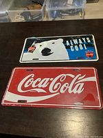 Photo showing two Coca-Cola license plates side by side, one with polar bear design, the other with Coca-Cola script logo.