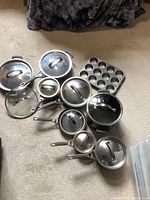 Overview of 8 Kirkland pots and pans with some glass lids, arranged on carpet, showing variety in sizes and lid types.