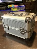 Full view of a silver steel makeup case with metal corner reinforcements, two front locks, and a handle on top.