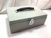 Photo of the closed cash box showing the textured light gray metal exterior, black handle, and chrome latch lock.