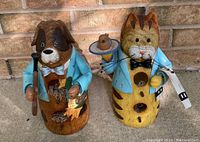 Photo of two decorative wooden bird houses, a dog and a cat figure both wearing blue jackets with multiple small nesting holes visible.