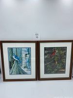 Two wood framed and matted bird pictures against a wall. Left picture features a blue jay in a forest scene; right picture features two cardinals.
