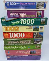 Stack of nine jigsaw puzzle boxes from various brands showing piece counts and some scenes on boxes.