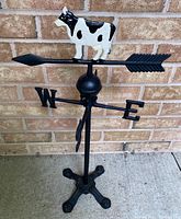 Full view of the cast iron cow weather vane on a stand placed in front of a brick wall, showing the black paint on the base and directional markers with the cow mounted on the arrow.