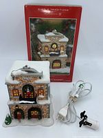 Porcelain fire station building with Christmas wreaths, snow accents, and plugged-in light cord with clip in front of original red branded box.