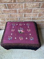 Overall view of the footstool showing the needlepoint top with floral pattern and dark border fabric on wooden frame.