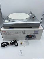 Full view of the silver Ion USB belt drive turntable placed on top of its original box with power cord and small accessory in front.