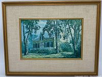 Full image of framed textured wall art depicting a stone house with trees in the foreground. The artwork is surrounded by a beige fabric mat and a wood frame with gold trim.