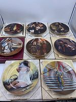 Nine collector plates displayed, each showing scenes from Americana and nostalgic moments, mainly by Norman Rockwell and including a certificate of authenticity with some water damage visible on boxes and papers.