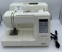 Front view of white Sears Kenmore sewing machine with control panel and stitch selection buttons visible. Hard shell carrying case in background and power cord laid beside machine.