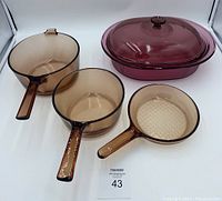 Three amber glass cookware pieces including two saucepans and one skillet alongside a cranberry glass oval casserole with lid