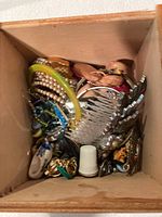 Overhead view inside wooden box showing various jewellery pieces piled together including bracelets, brooches, combs, and small decorative items.