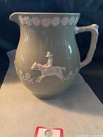 Front view of vintage Spode pitcher showing embossed horse and rider and floral relief pattern on sage green ceramic body with branch style handle.