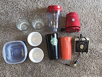 All kitchen and drinkware items arranged on carpet including Starbucks tumblers, blender parts, side bowls, Tupperware container, glass cups, and alarm clock.