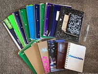 Top-down image showing multiple spiral-bound notebooks in different colors, several bound notebooks, a black and white composition book, and a white notebook with #launchlife on the cover spaced out on a carpeted floor.