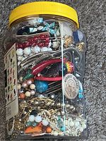 Plastic jar filled with assorted jewelry items including chains, beads, and earrings, showing the overall jar and contents layered.