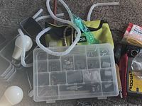Photo showing plastic storage organizer with small compartments containing various small metal hardware pieces, coiled white tubing, white funnel, and a yellow tool bag.