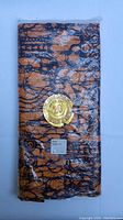 Folded batik sarong packaged in plastic wrap with a circular gold and red sticker and product label visible.