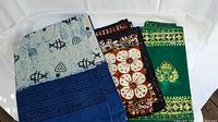 Four handmade Balinese batik textiles folded and layered, featuring blue and white fish pattern, brown floral pattern, and green geometric and lotus pattern.