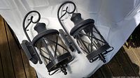 Pair of black metal outdoor sconce light fixtures with curved decorative hooks and glass panel lantern bodies on white background outside.