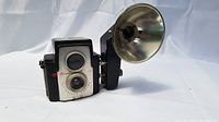 Side angled view showing the camera body with brown sides, dual lens faceplate, attached large flash reflector, and neck strap.